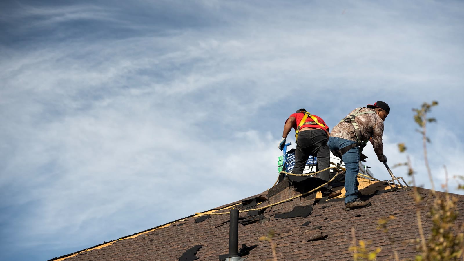 Two roofers working on an asphalt shingle roof under a wide sky after a hail storm